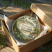 A decorative ornament in a box, featuring a scenic landscape with a lighthouse and a coastal area.