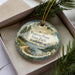 A decorative ornament with a scenic landscape of Penrose Point State Park in Washington, featuring pine trees, a river, and mountains.