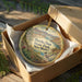 A decorative ornament in a box, featuring a scenic landscape with a bird and a bridge.