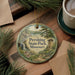 A round ornament with a scenic landscape of Pershing State Park, Missouri, featuring a waterfall and pine trees, is displayed on a wooden surface alongside a cup of coffee and some papers.