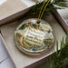 A decorative ornament in a box, featuring a scenic landscape with mountains and pine trees.