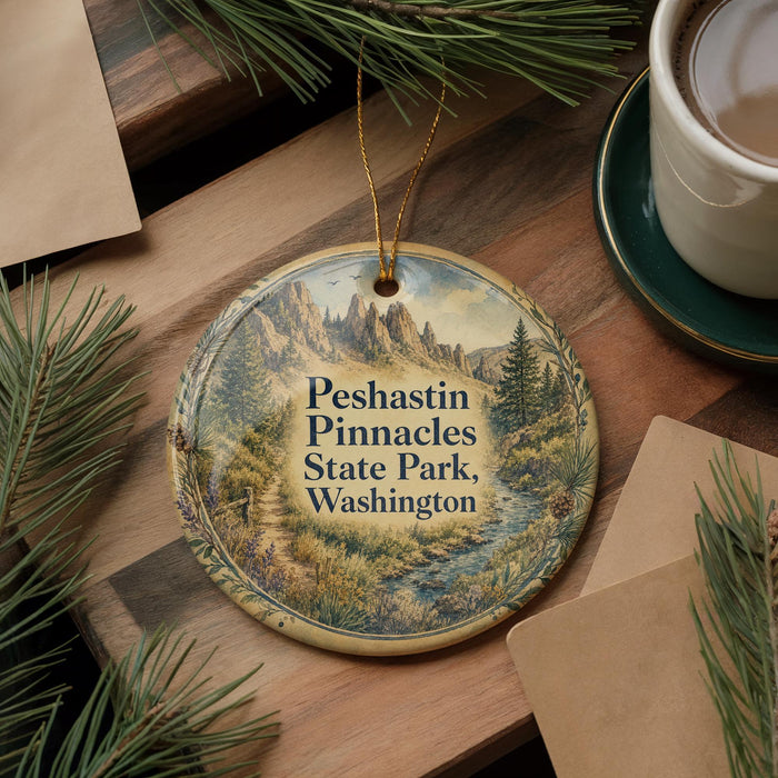 A decorative ornament featuring a scenic landscape of mountains and pine trees, with the text "Peshastin Pinnacles State Park, Washington" printed on it. The ornament is hanging on a wooden surface, surrounded by pine branches and a cup of coffee.