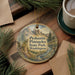A decorative ornament featuring a scenic landscape of mountains and pine trees, with the text "Peshastin Pinnacles State Park, Washington" printed on it. The ornament is hanging on a wooden surface, surrounded by pine branches and a cup of coffee.