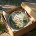 A decorative ornament in a box, featuring a scenic landscape with a wooden walkway and a house, surrounded by greenery.