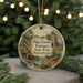 A round ornament with a painting of a scenic landscape, featuring a chimney, a mill, and a river, hanging in front of a blurred background with Christmas lights.