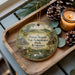 A wooden tray with a decorative ornament featuring a scenic landscape and wildlife, including a deer, a bird, and a house, along with pine cones and a lit candle.