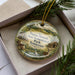 A decorative ornament with a landscape scene featuring pine trees, a house, and wildlife, including a bird, is displayed in a box.