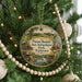 A round ornament hanging on a Christmas tree, featuring a scenic landscape with a house, trees, and a bird.