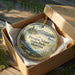 A decorative ornament in a wooden box, featuring a scenic landscape with mountains, trees, and a lake.