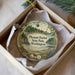A decorative ornament in a box, featuring a scenic landscape with a lighthouse, trees, and a body of water.