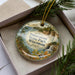 A decorative ornament in a box, featuring a scenic landscape with a lake, mountains, and pine trees.