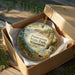 A decorative ornament in a wooden box, featuring a scenic landscape with a waterfall and forest.