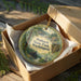 A decorative ornament in a box, featuring a scenic landscape with a cabin and trees, with the text "Potlatch State Park, Washington" printed on it.