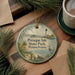A round ornament with a lighthouse and a scenic view of Presque Isle State Park, Pennsylvania, is displayed on a wooden surface alongside a cup of coffee and some pine branches.