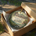 A decorative ornament in a wooden box, featuring an illustration of a fort surrounded by trees and a stream, with the text "Prickett's Fort State Park, West Virginia" printed on it.