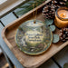 A decorative ornament featuring a scenic landscape of Prince Gallitzin State Park, Pennsylvania, with a candle and pine cones on a wooden tray.