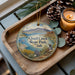 A decorative ornament featuring a scenic image of Quail Creek State Park in Utah, with pine cones and a lit candle on a wooden tray.