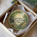 A decorative ornament in a box, featuring a scenic landscape with a house, trees, and a bird.