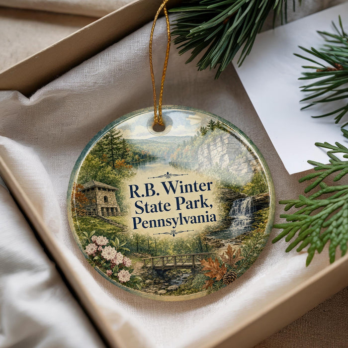 A decorative ornament with a scenic landscape of Pennsylvania, featuring a house, trees, and a waterfall, is displayed in a box with pine branches.