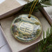 A decorative ornament in a box, featuring a scenic landscape with mountains, forests, and a waterfall.