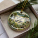 A decorative ornament with a scenic image of Raymond Winter State Park in Pennsylvania, featuring a waterfall, pine trees, and a cabin.