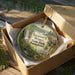 A round ornament in a box, featuring a scenic image of Raymond B Winter State Park in Pennsylvania.