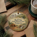 A decorative ornament with a painting of a cabin and a waterfall, surrounded by pine branches and a cup of coffee on a wooden surface.