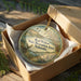 A decorative ornament in a wooden box, featuring a scenic landscape with a river, mountains, and wildlife.
