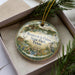 A decorative ornament in a box, featuring a landscape scene with a bridge, birds, and other natural elements.
