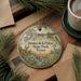 A decorative ornament with a scenic landscape of a lake, palm trees, and wildlife, including a deer, is displayed on a wooden surface alongside a cup of coffee and some papers.