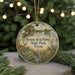 A round ornament with a scenic landscape featuring a lake, palm trees, and wildlife, including a bird and a deer, is displayed on a table with a blurred background of lights.
