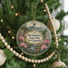 A round ornament hanging on a Christmas tree, featuring a scenic landscape of mountains, pine trees, and pink flowers.
