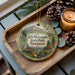 A decorative ornament featuring a scenic image of Rib Mountain State Park, Wisconsin, with pine cones and a lit candle on a wooden tray.