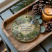 A decorative ornament featuring a scenic image of Ridley Creek State Park, Pennsylvania, with pine cones and a lit candle on a wooden tray.