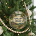 A round ornament hanging on a Christmas tree, featuring a scenic landscape with a house, trees, and a waterfall.