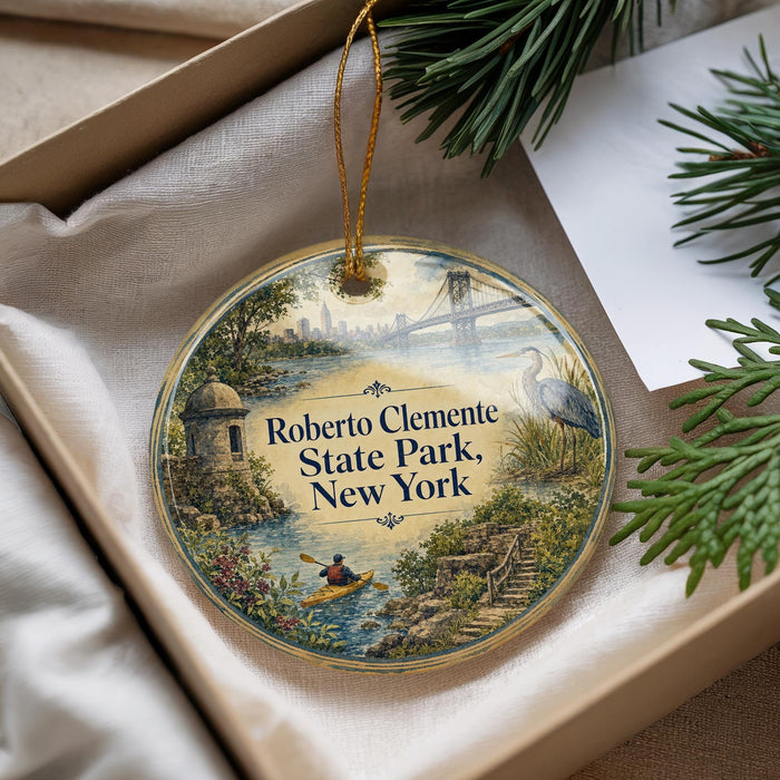 A decorative ornament with a scenic image of a park, featuring a person kayaking, a bridge, and a bird, is displayed in a box with pine branches.