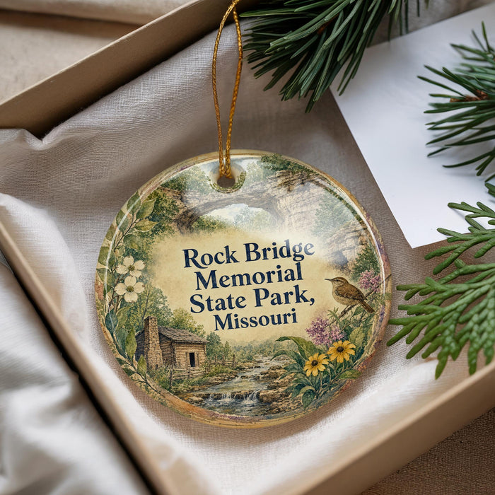 A decorative ornament with a scenic landscape of a river, cabin, and flowers, hanging from a string.