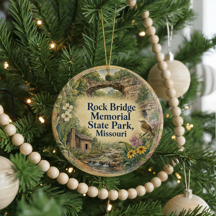A round ornament hanging on a Christmas tree, featuring a scenic landscape with a cabin and a bird.