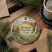 A decorative ornament with a scenic image of Rockport State Park, Utah, featuring a house, a waterfall, and a boat on a lake. The ornament is hanging on a wooden surface, surrounded by pine branches and a cup of coffee.