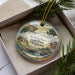 A decorative ornament in a box, featuring a scenic landscape with a waterfall and a house, with the text "Roman Nose State Park, Oklahoma" prominently displayed.
