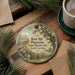 A round ornament with a painting of a house and a tree, surrounded by pine branches, sits on a wooden surface.