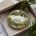 A decorative ornament in a box, featuring a scenic landscape with a house, trees, and a waterfall.
