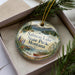 A decorative ornament in a box, featuring a scenic landscape with mountains, pine trees, and a lake.