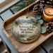 A decorative ornament with a scenic landscape of Salmon Lake State Park, Montana, featuring a lake, mountains, pine trees, and a canoe. The ornament is displayed on a wooden tray, accompanied by pine cones and a lit candle.