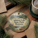 A decorative ornament with a scenic image of Salmon Lake State Park, Montana, featuring a lake, mountains, and pine trees. The ornament is hanging on a wooden surface, surrounded by pine branches and a cup of coffee.