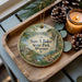 A decorative ornament featuring a scenic landscape with a waterfall, pine cones, and a candle on a wooden tray.
