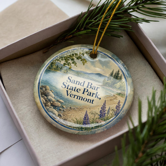A decorative ornament in a box, featuring a scenic landscape with a lake, mountains, and pine trees.