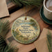 A round ornament with a scenic illustration of a deer in a field, surrounded by pine branches, sits on a wooden surface next to a cup of coffee.