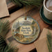 A decorative ornament featuring a scenic landscape with mountains, pine trees, and a cabin, hanging on a wooden surface.