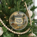 A round ornament hanging on a Christmas tree, featuring a scenic landscape with mountains, a cabin, and a lake.